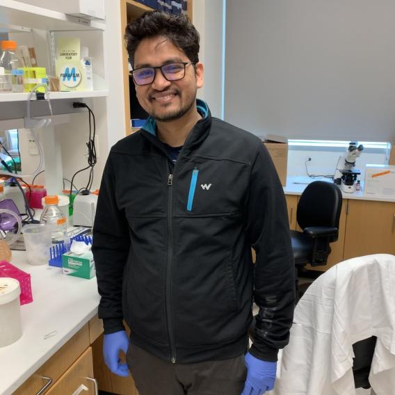 Smiling man with dark hair and trim facial hair, wearing black clothes, dark-rimmed glasses, and blue latex gloves, standing in a lab.