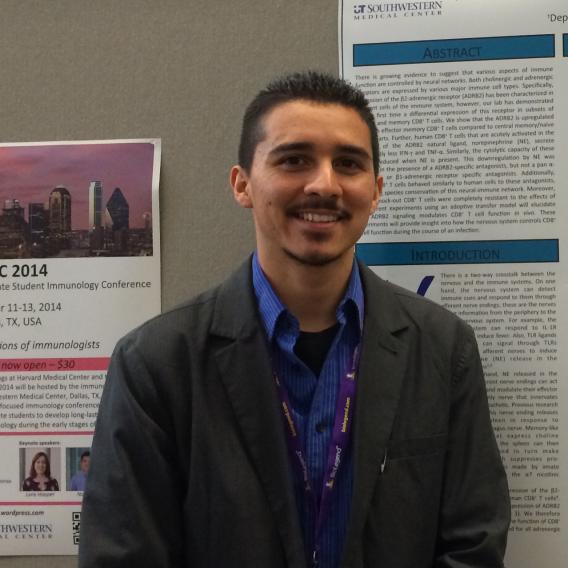 Dr. Estrada, with trim dark hair, beard and mustache, standing in front of a bulletin board, wearing a dark gray suit jacket over a blue shirt.