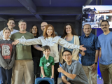 Smiling woman with red hair, wearing a white blouse and black pants, stands with arms spread amid a group of 6 men, 2 women, a boy, and a girl.